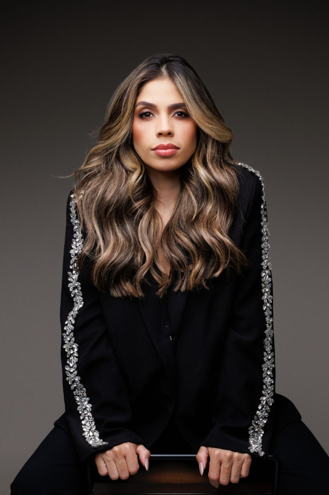Bruna Kimberlly sentada, de blazer preto com pedraria, com o cabelo longo em ondas