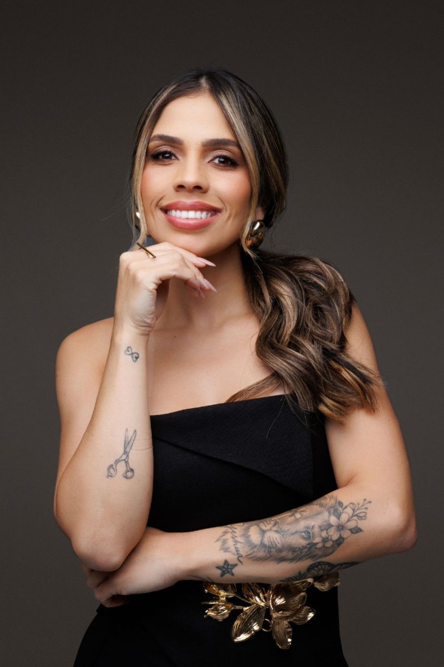 Bruna Kimberlly sorrindo, com a mão no queixo, de preto com cinto dourado